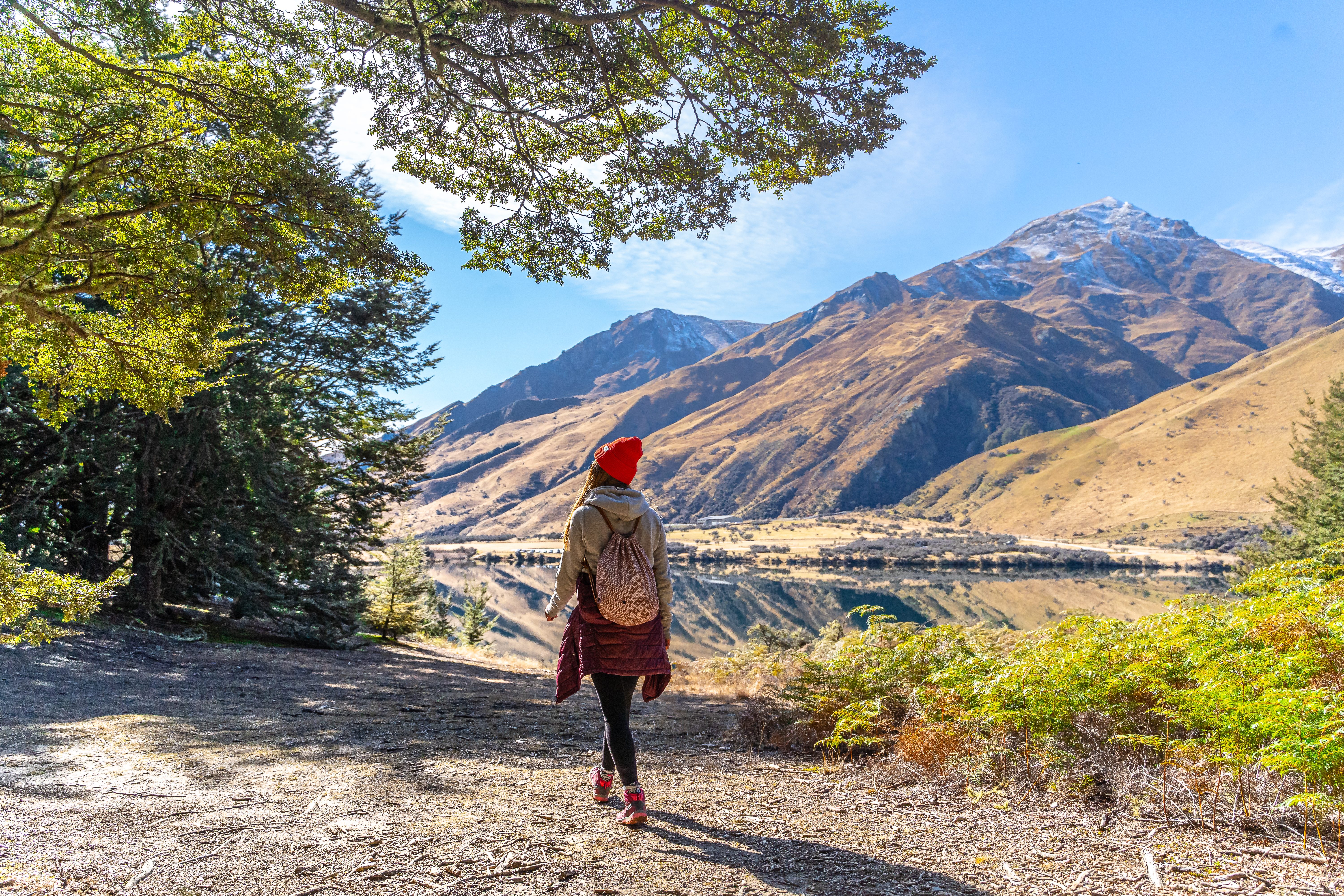 A person in red hat hiking by Moke Lake, surrounded by mountains and trees under a clear blue sky.
