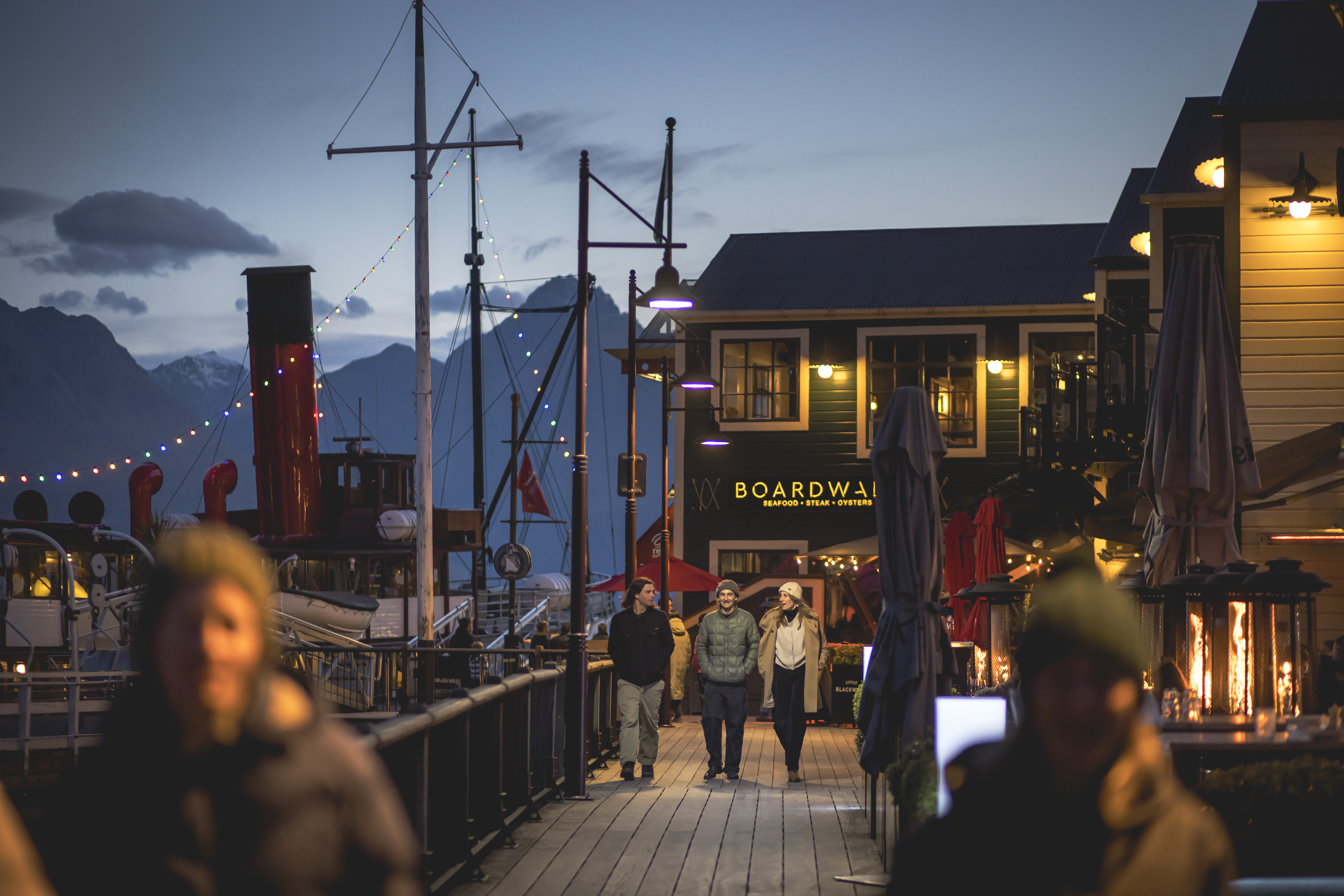 People walking on a Queenstown boardwalk at dusk, surrounded by quaint shops and a mountainous backdrop.