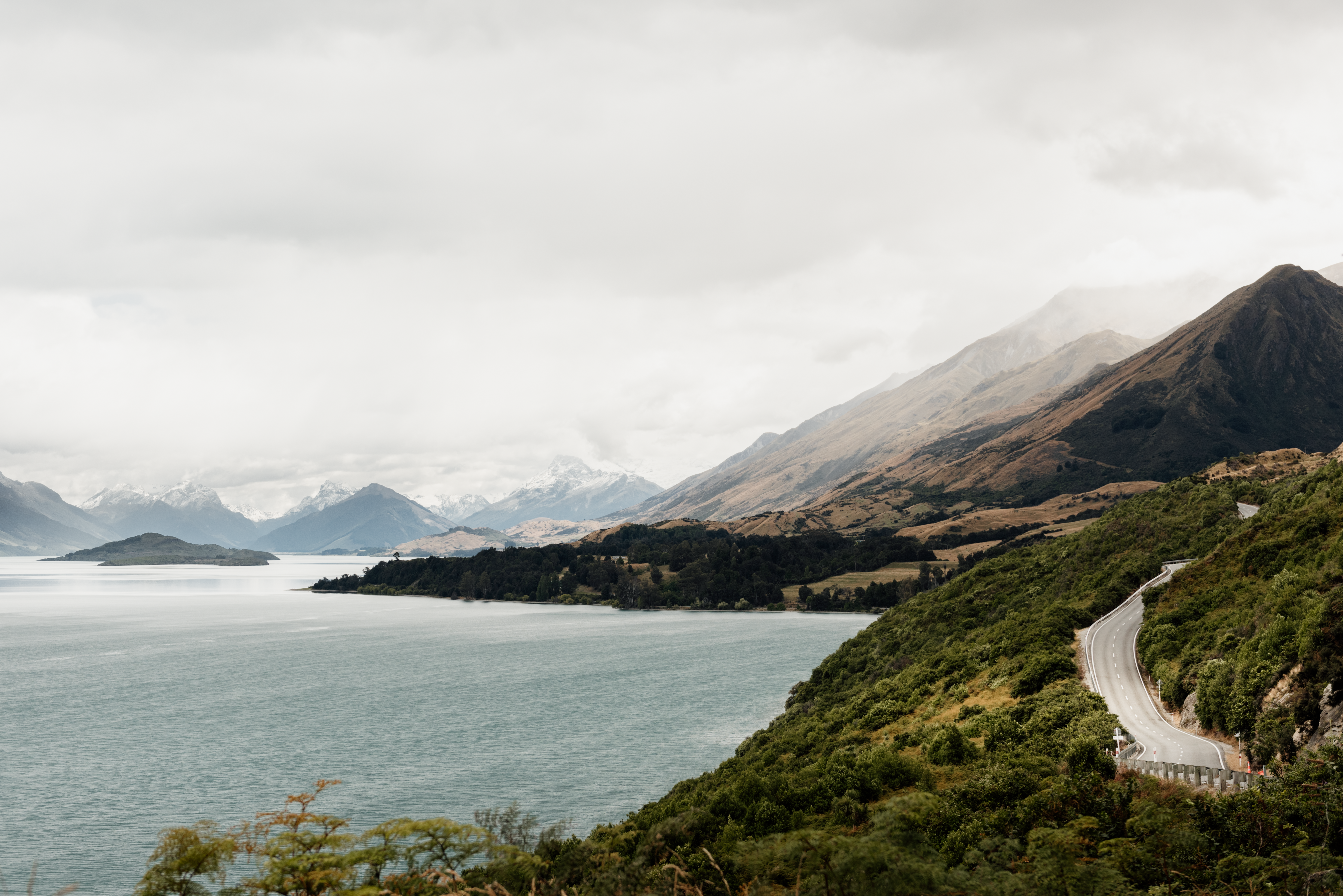 Glenorchy Road along a lake with mountains in the background under a cloudy sky.