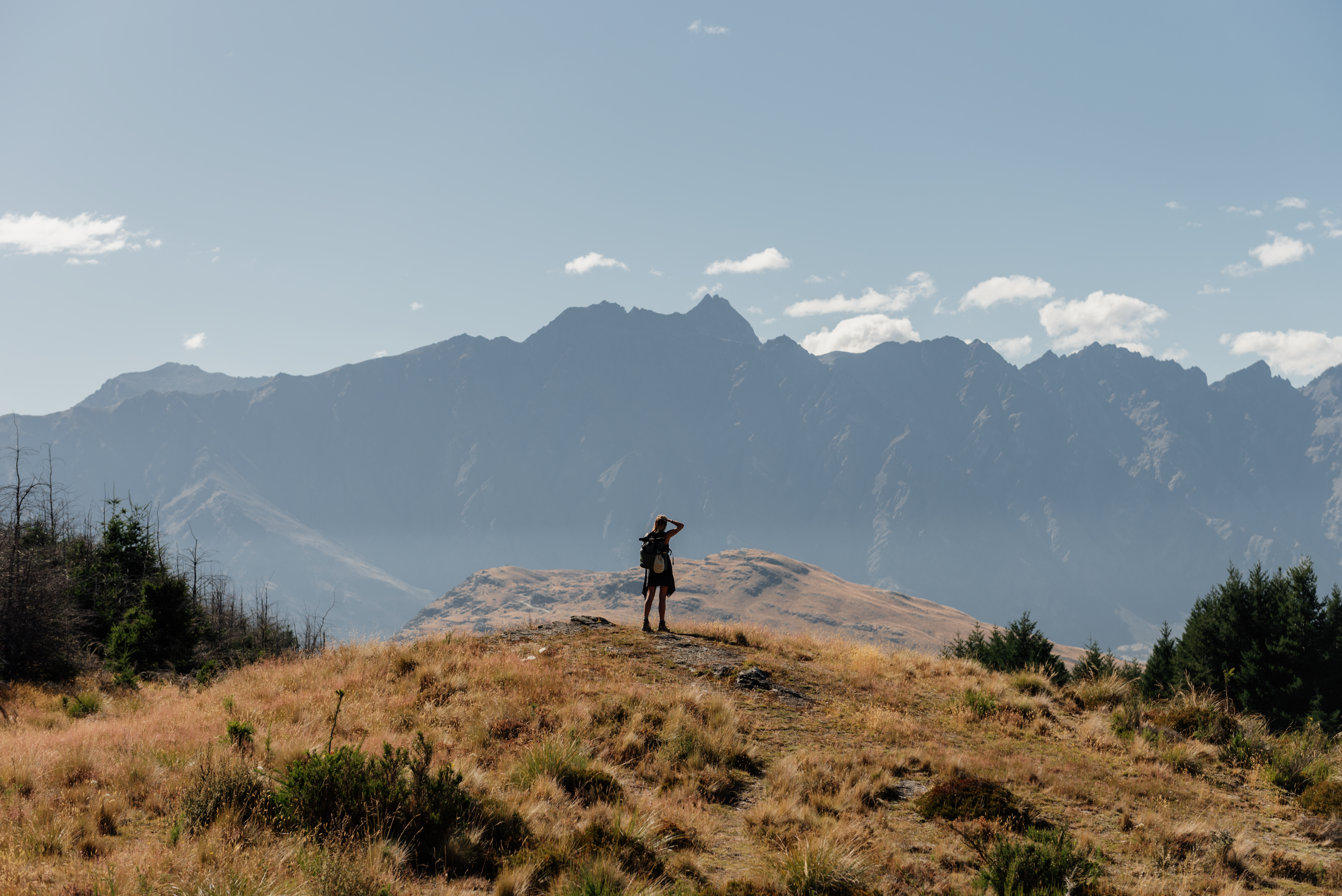A person standing on a grassy hilltop, surrounded by mountains under a clear sky.