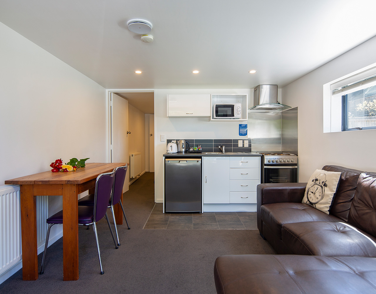 A compact kitchen and living area in a one-bedroom apartment at Pinewood with dining table, brown leather sofa, and modern appliances.
