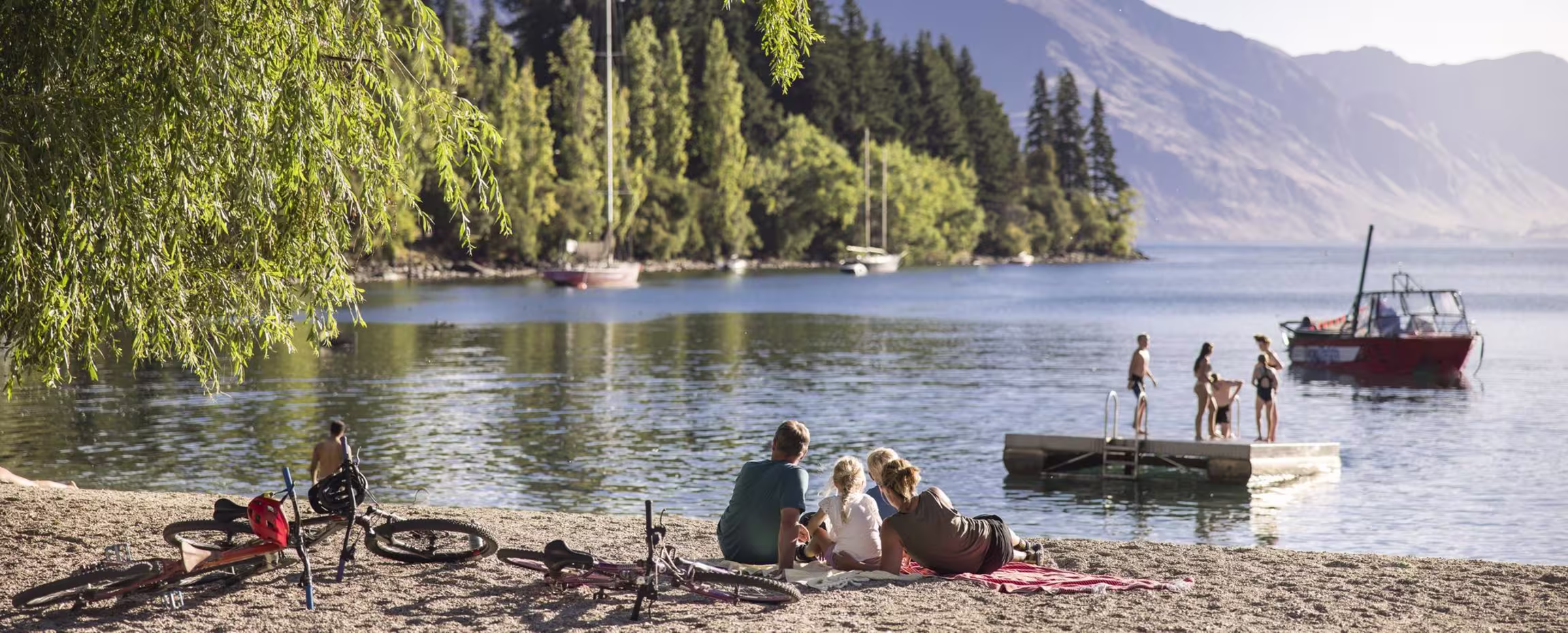 people relaxing by a calm lake with a small boat and forested mountains in the background